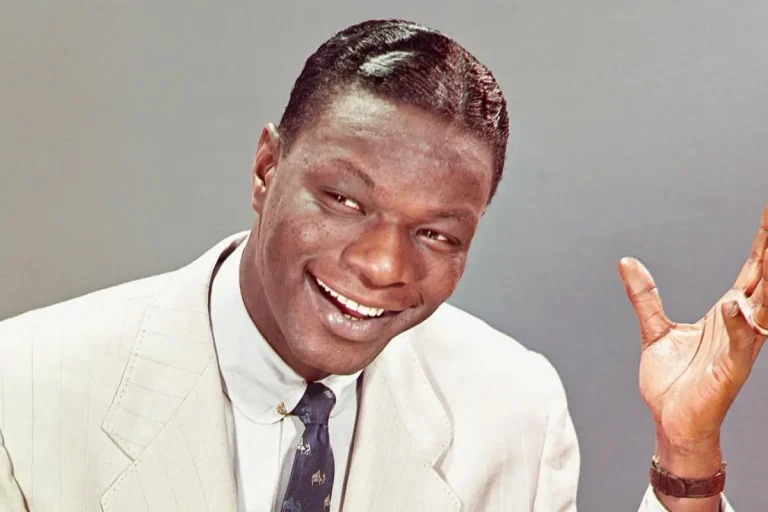 Nat King Cole in a light-colored suit, smiling broadly and raising one hand.