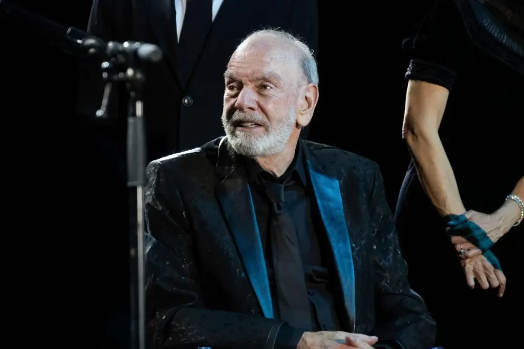 Sweet Caroline: The Strange, Sweet Story Behind Neil Diamond’s Anthem Neil Diamond seated on stage in a black suit with a blue lapel.