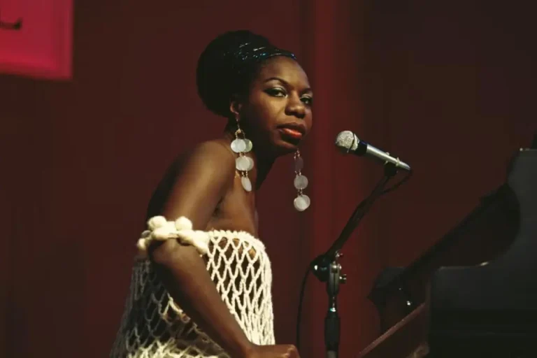 Nina Simone a netted dress and large dangling earrings performs at a piano, turning toward the microphone under stage lighting.