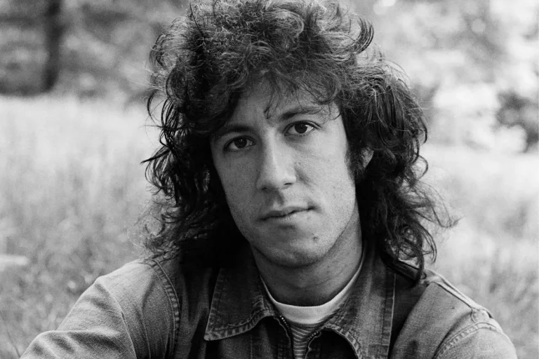 Peter Green gazes softly at the camera while sitting outdoors in a grassy, blurred background.