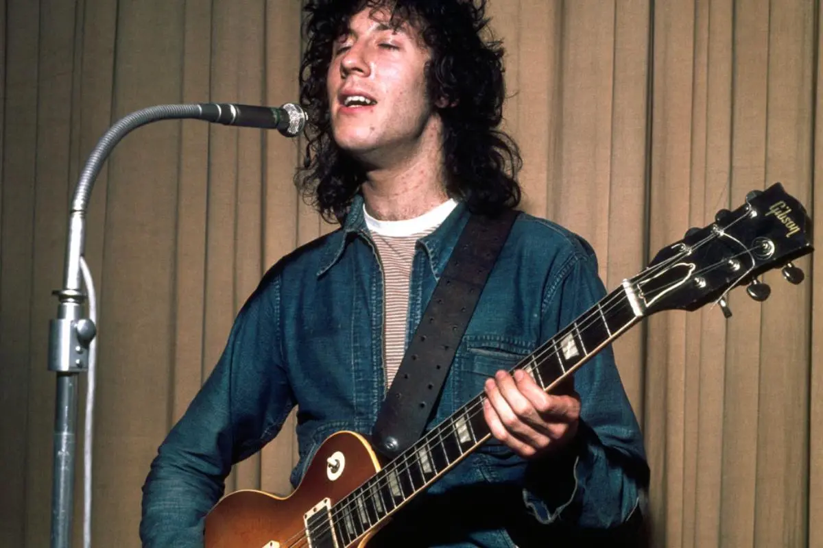 Peter Green sings into a microphone while playing a Gibson guitar, eyes closed in concentration.