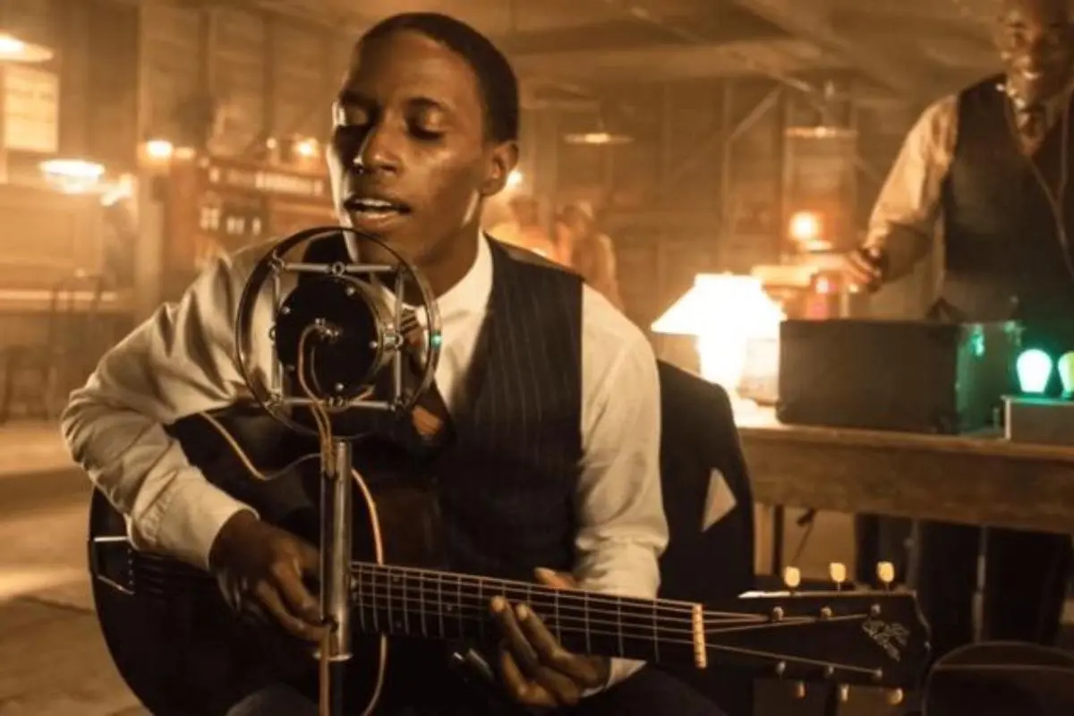 Robert Johnsons sings into a vintage microphone while playing an acoustic guitar in a dimly lit recording space.