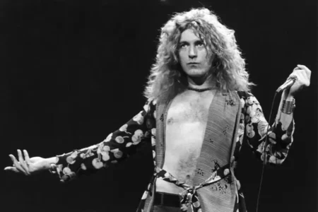 Robert Plant with long curly hair performs onstage, holding a microphone and wearing an open patterned robe.