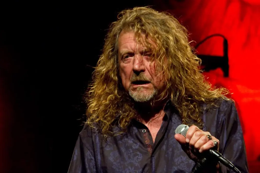 Robert Plant holds a microphone and sings intensely under warm red stage lighting.