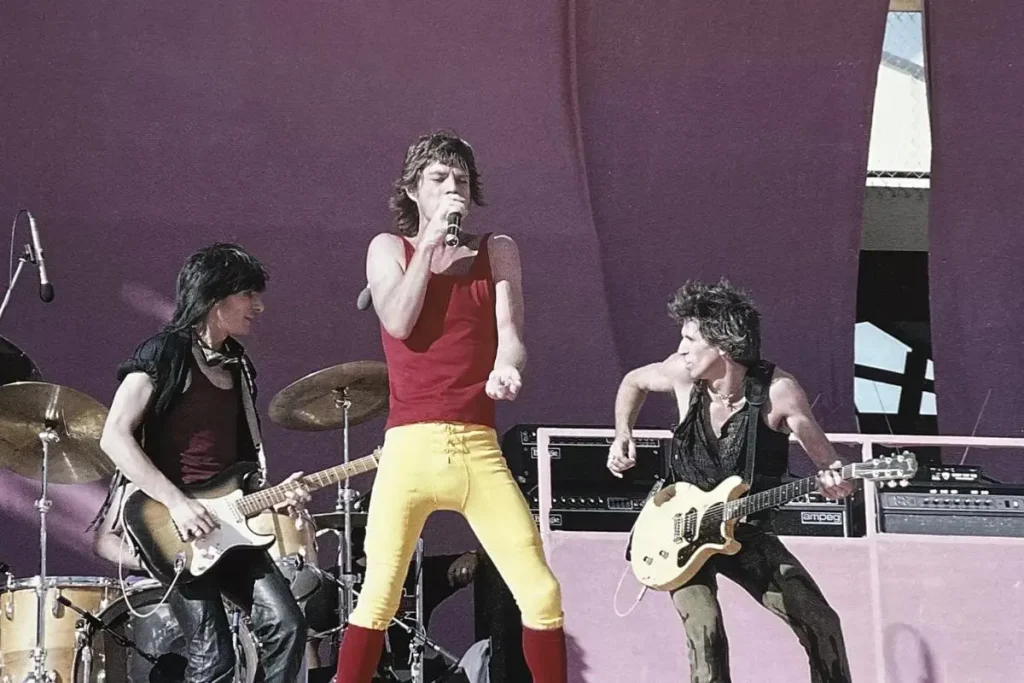 Mick Jagger in a red tank top and yellow pants sings onstage.