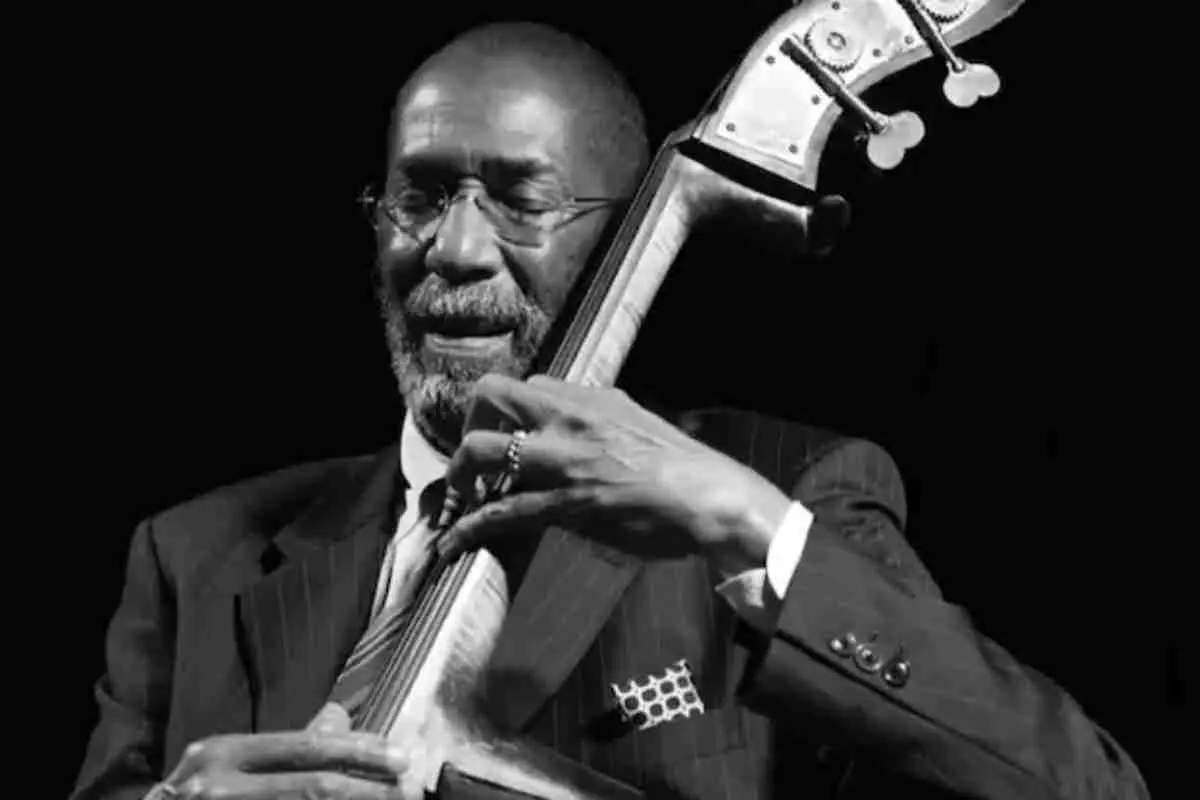 Ron Carter playing the upright bass in a black-and-white performance photo, wearing a suit and tie, focused on the instrument against a dark background.