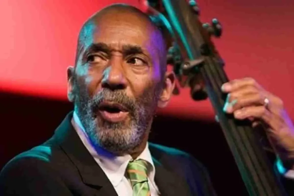 Ron Carter performing on stage with an upright bass, wearing a dark suit and green tie, captured mid-performance under colorful stage lighting.