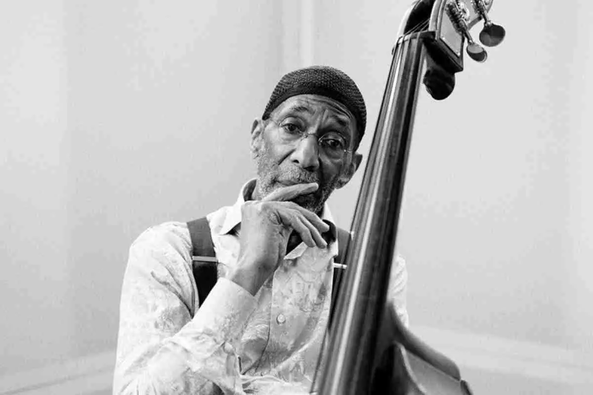 Ron Carter posing in a black-and-white portrait beside an upright bass, resting his hand near his face, wearing a patterned shirt, suspenders, and a knit cap with a contemplative expression.