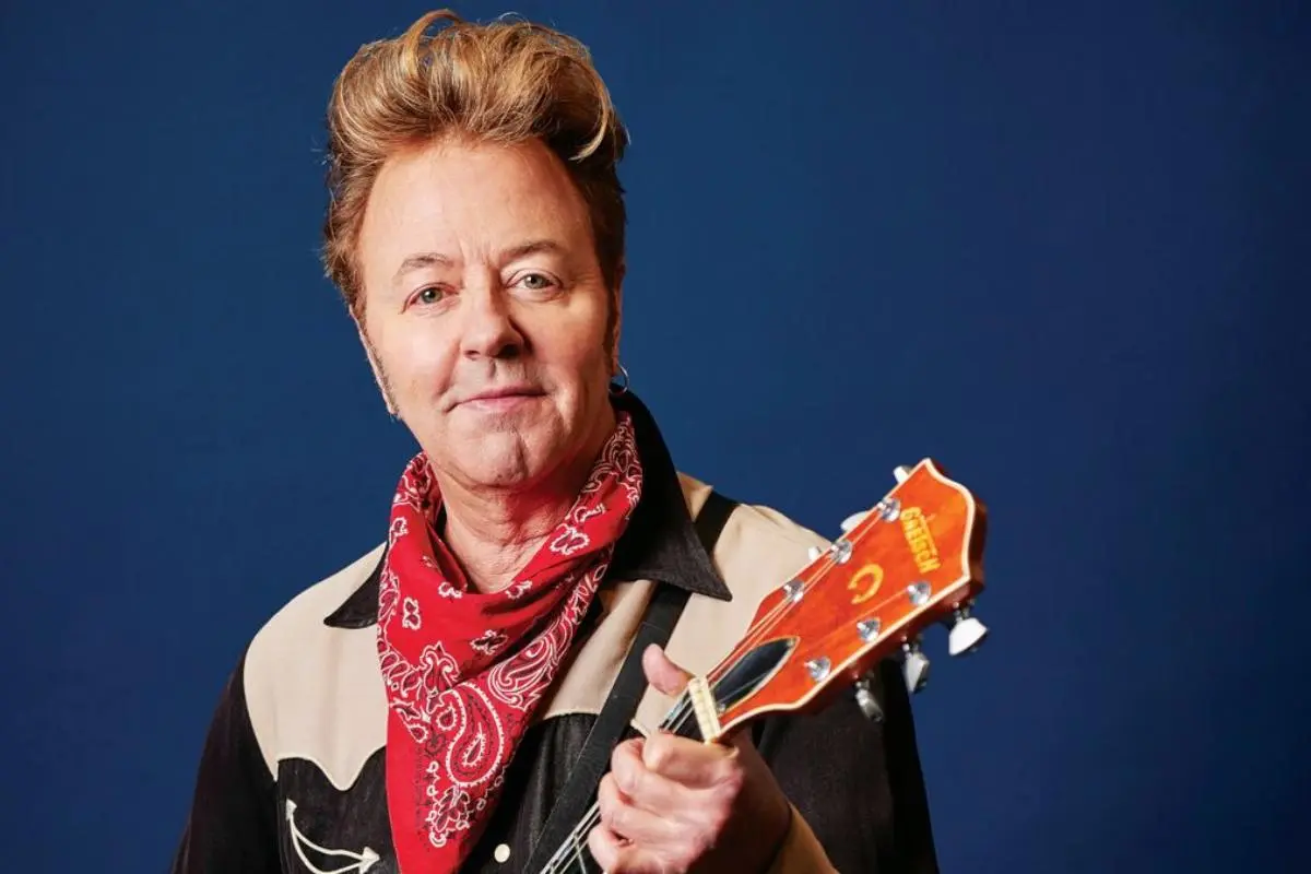 Portrait of Brian Setzer holding a guitar against a blue background.