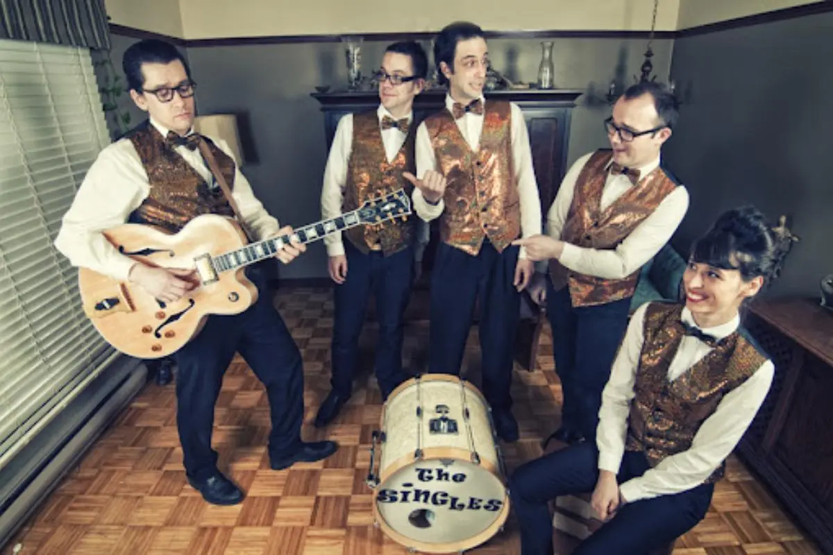 Single Band wearing matching vests and bow ties, posing indoors with guitars and a bass drum.
