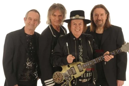 The Slade Band poses against a white background, smiling as one member holds a sparkling guitar.