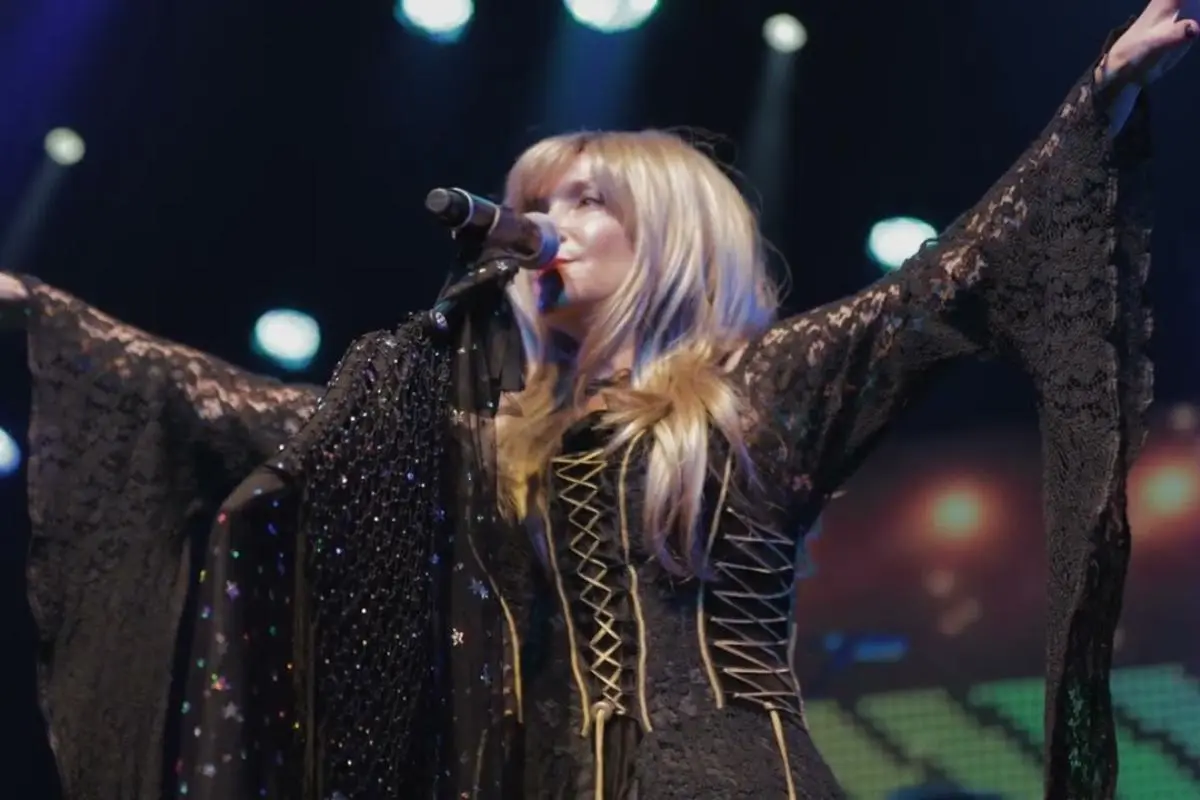 Stevie Nicks sings into a microphone with arms outstretched, wearing a black lace outfit on a lit stage.