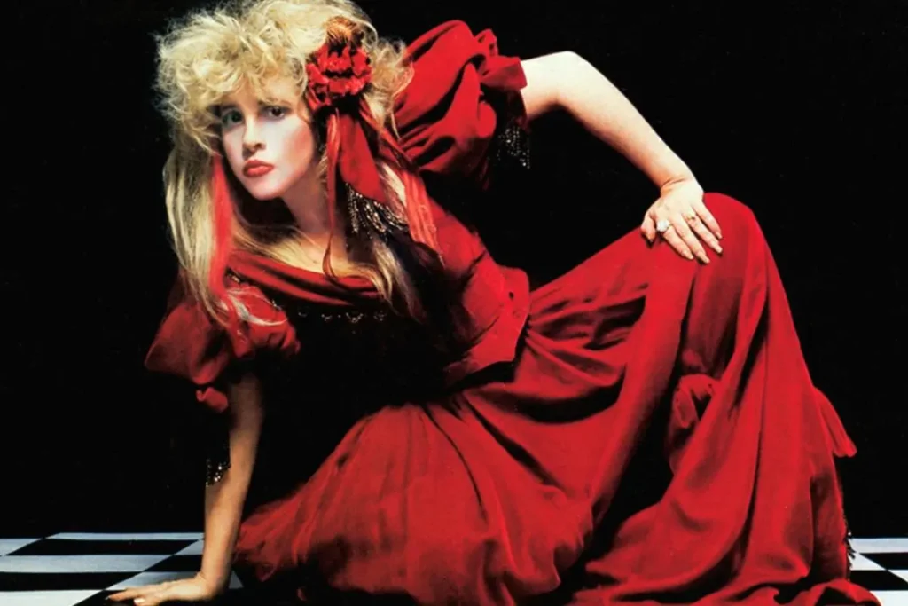 Stevie Nicks in a dramatic red dress poses against a dark background, styled in a vintage, theatrical look.