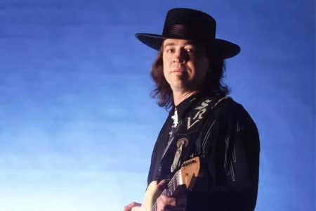 Stevie Ray Vaughan standing with a Fender Stratocaster against a blue gradient background.