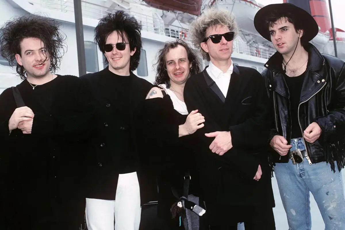The Cure band members posing together outdoors, dressed in black and casual attire.
