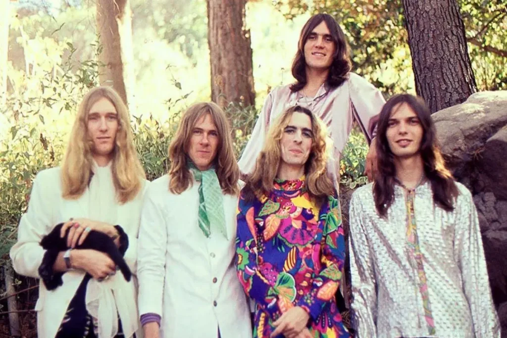 Alice Cooper band stand among trees and rocks, dressed in colorful and ornate clothing, with soft natural light illuminating the group.