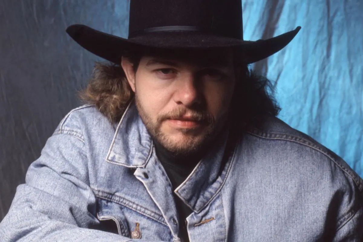 Toby Keith with long hair and a beard wears a black cowboy hat and denim jacket, looking directly at the camera.