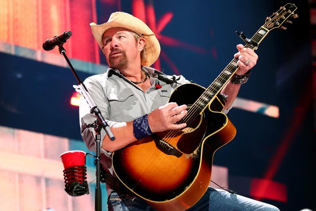 Toby Keith wearing a cowboy hat strums an acoustic guitar during a live concert.