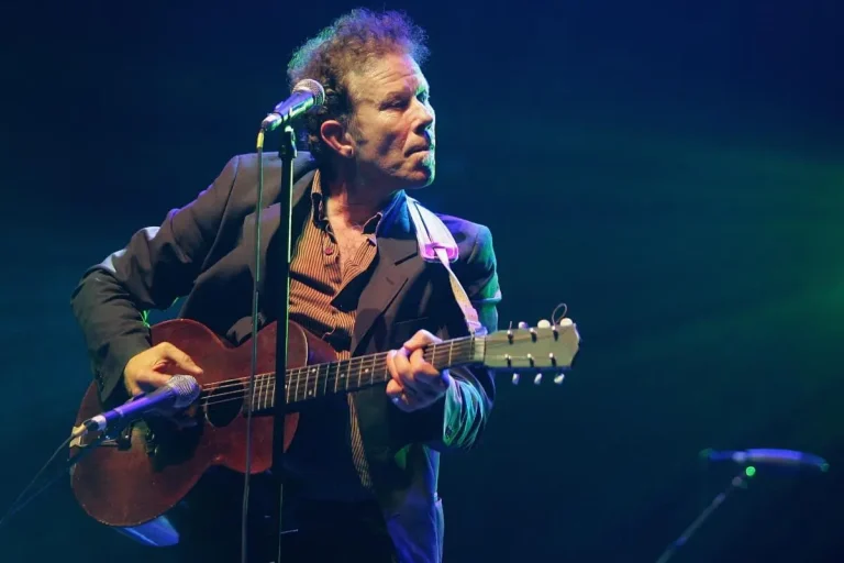 Tom Waits performs onstage with an acoustic guitar, leaning into the microphone under moody blue-green lighting.