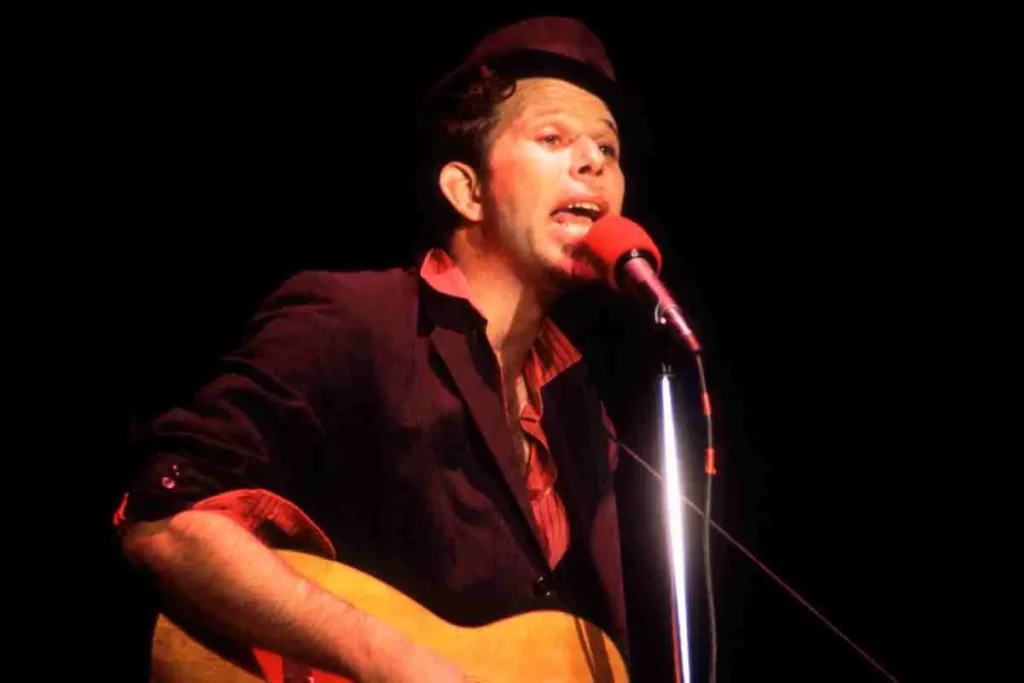 Tom Waits performing on stage, singing into a microphone while playing an acoustic guitar, wearing a dark jacket and hat under warm red stage lighting.