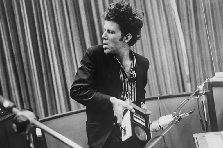 Tom Waits in a recording studio, mid-motion, holding a portable tape recorder.