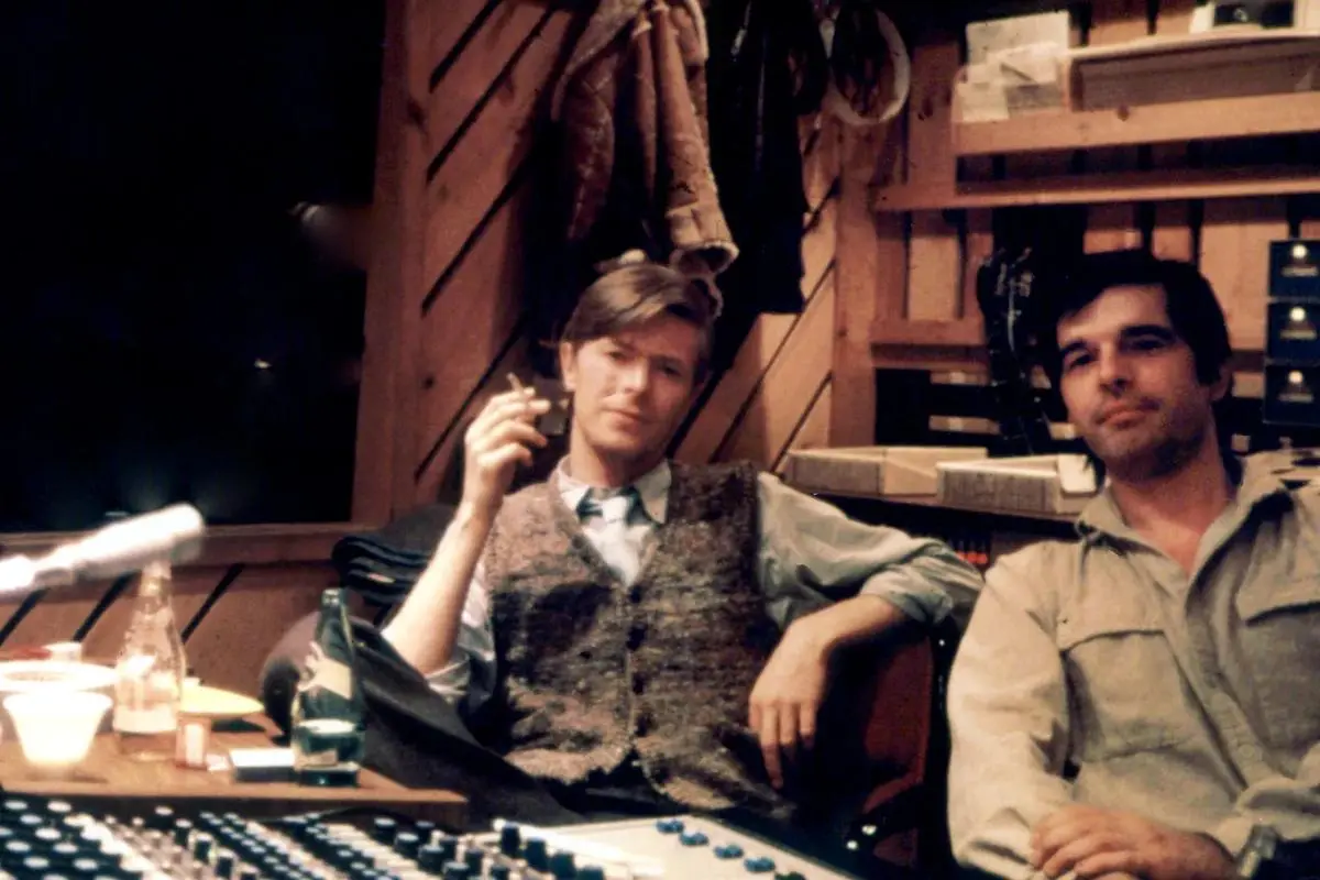 Tony Visconti with other man sitting in a recording studio surrounded by equipment, with one smoking a cigarette as they relax between sessions.