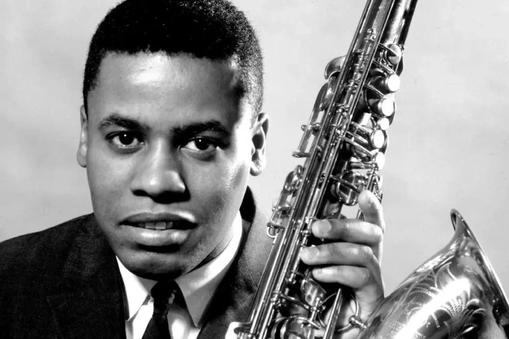 Wayne Shorter wearing a suit and tie, holding a saxophone upright beside his face while looking directly at the camera.