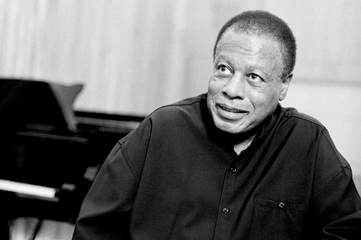 Wayne Shorter sitting near a piano, wearing a dark button-up shirt.
