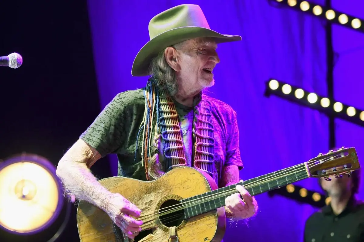 Willie Nelson plays his famously weathered acoustic guitar onstage under purple lighting.