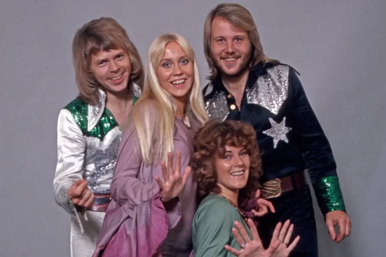 ABBA posing together in colorful 1970s stage outfits, smiling and gesturing toward the camera during a promotional photo shoot.