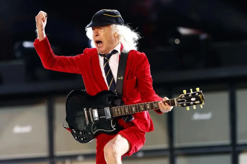 ACDC jumps onstage while playing a black electric guitar, raising a fist mid-performance under stadium lights.