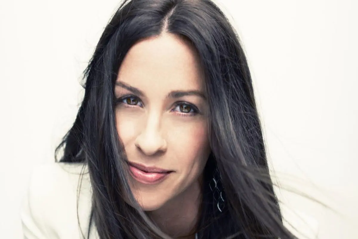 Alanis Morissette portrait with long dark hair, softly smiling against a light background.