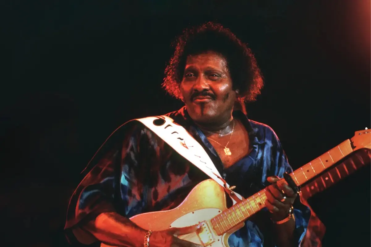 Onstage beneath warm red lighting, the guitarist smiles confidently while working the fretboard, embodying the cool authority and sharp precision of his live performances.