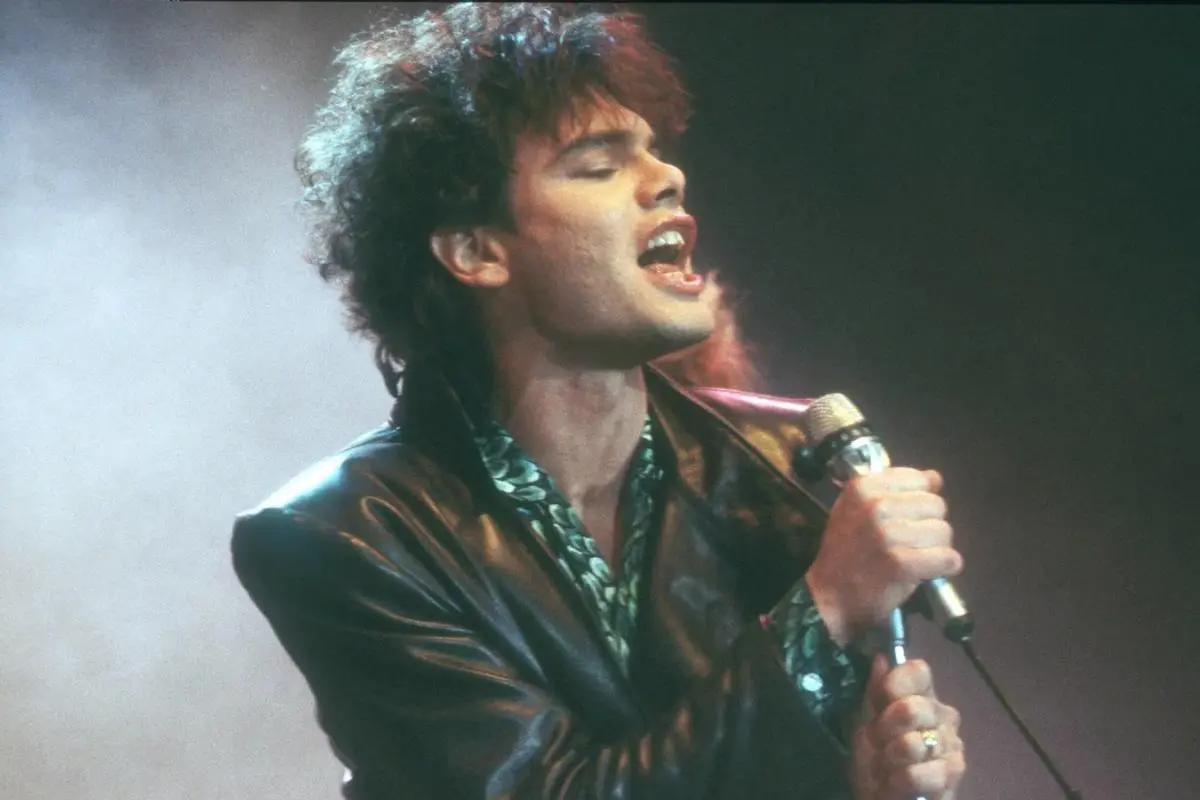 Alphaville performing on stage, eyes closed, singing into a handheld microphone while wearing a leather jacket under dramatic stage lighting.