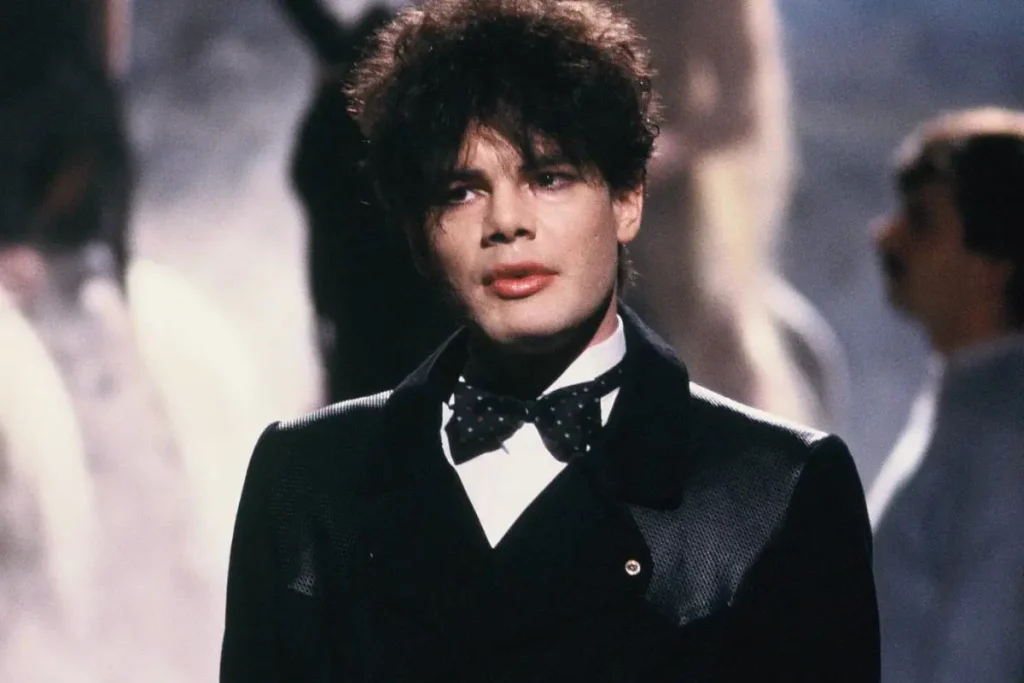 Alphaville wearing a formal black suit and bow tie, standing under soft stage lighting with a composed, reflective expression.