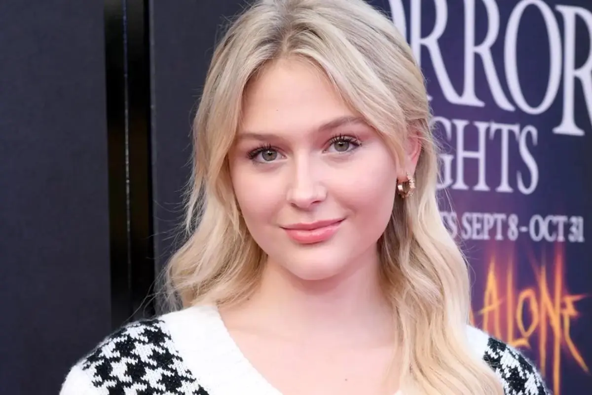 Alyvia Alyn Lind softly waved hair and natural makeup, wearing small earrings and a patterned top, smiling slightly at a media event backdrop.