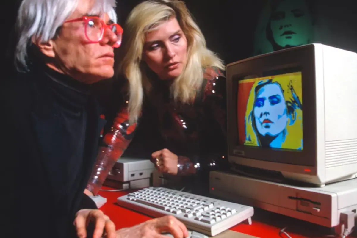 Andy-Warhol and Debbie Harry studying a digital portrait displayed on an early personal computer, highlighting experimentation at the intersection of technology and pop art.