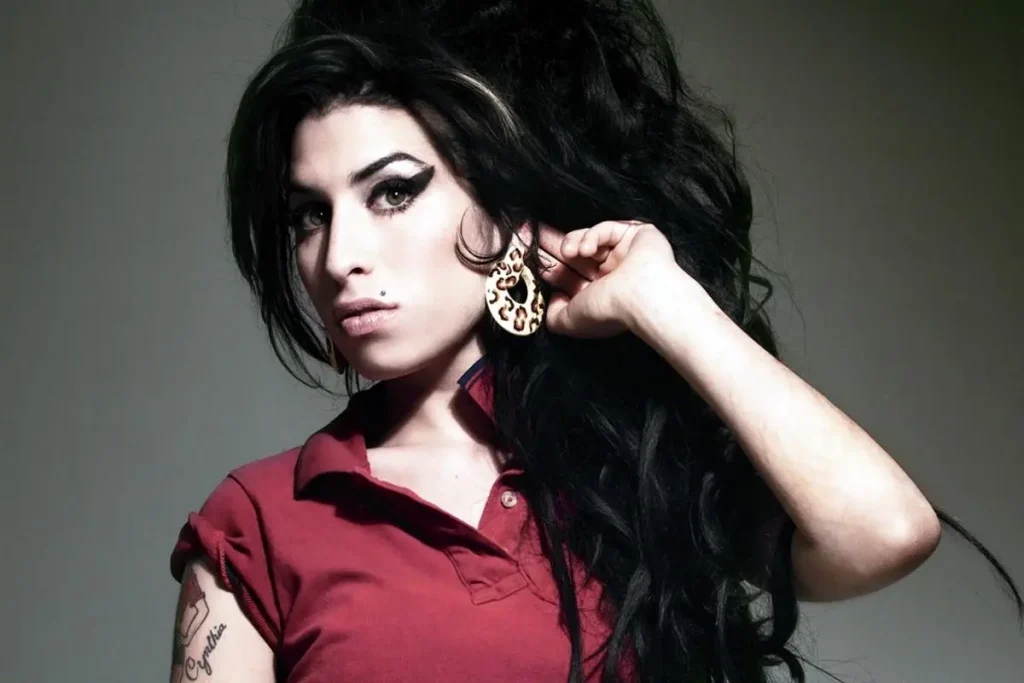 Amy Winehouse wearing a red collared shirt and large patterned earrings, looking directly at the camera.