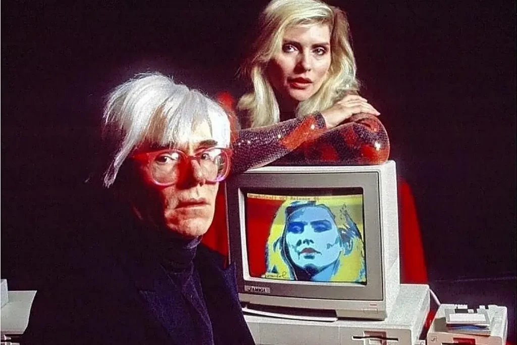 Andy-Warhol and Debbie Harry posed beside a computer showing a stylized digital portrait, emphasizing early digital creativity and the blending of art and emerging technology.