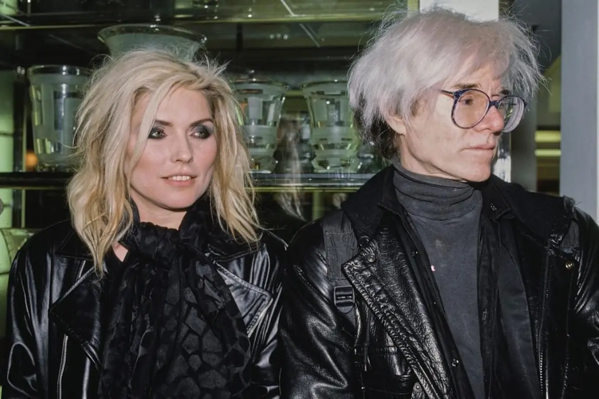 Andy-Warhol and Debbie Harry standing side by side in leather jackets, conveying cool detachment and the crossover of art, fashion, and music scenes.