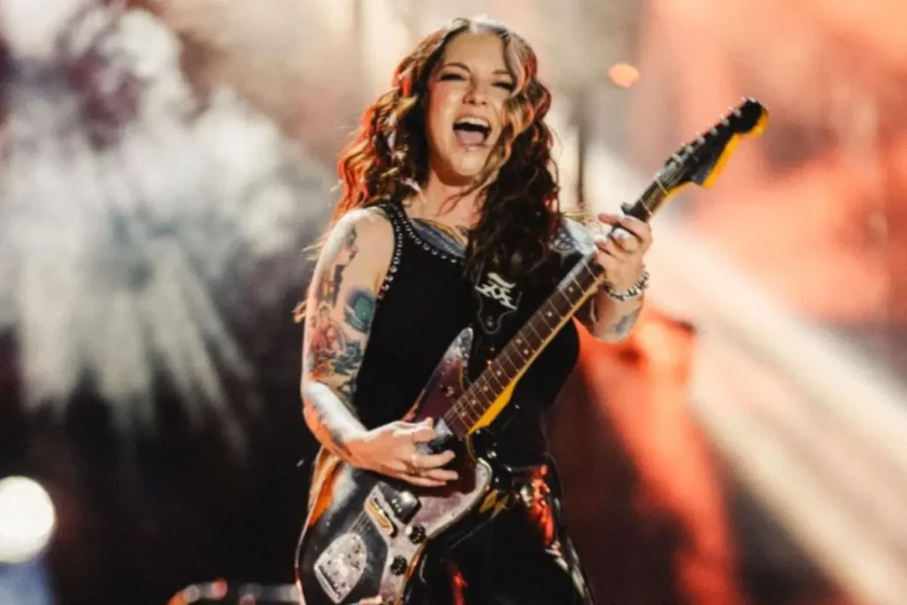 Ashley McBryde performing live on stage, singing and playing electric guitar, with tattoos visible on her arms.