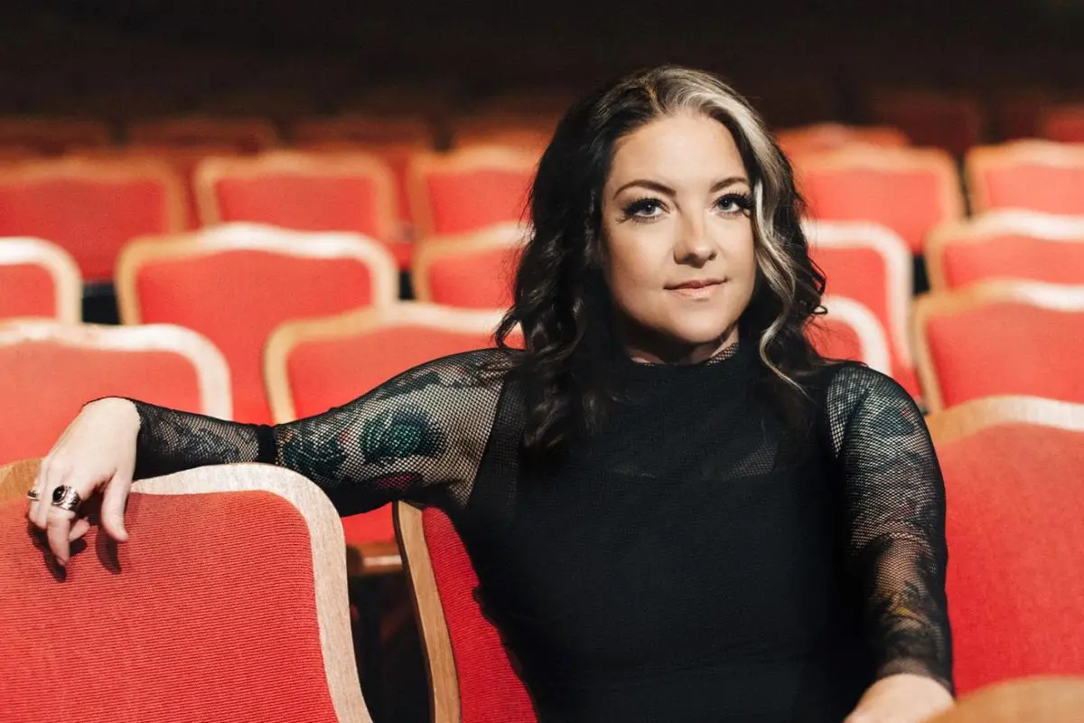 Ashley McBryde seated in a theater with red seats, wearing a black top and looking confidently toward the camera.