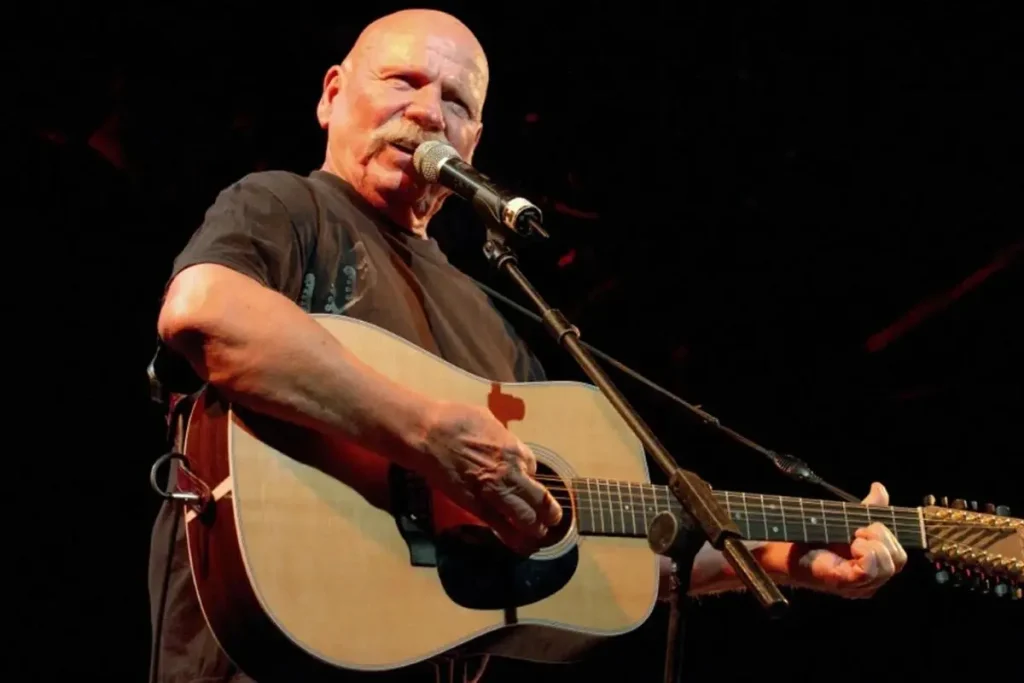 Barry McGuire singing into a microphone while playing an acoustic guitar under warm stage lighting.