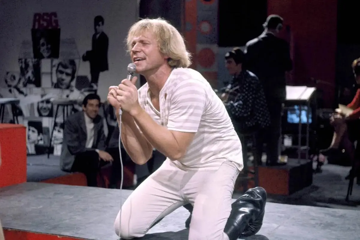 Barry McGuire on stage holding a microphone with both hands, performing passionately.