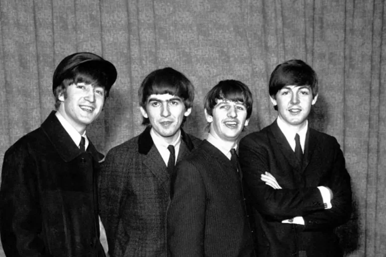 The Beatles—John Lennon, George Harrison, Ringo Starr, and Paul McCartney—standing side by side in matching suits, smiling slightly during an early 1960s press photo.
