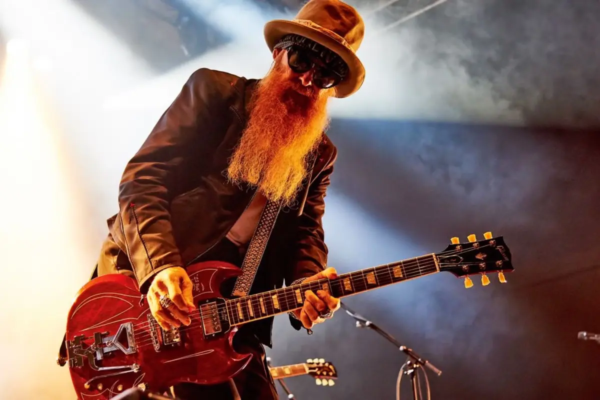 Billy Gibbons performs on stage, wearing a hat and sunglasses while playing an electric guitar under dramatic concert lighting.