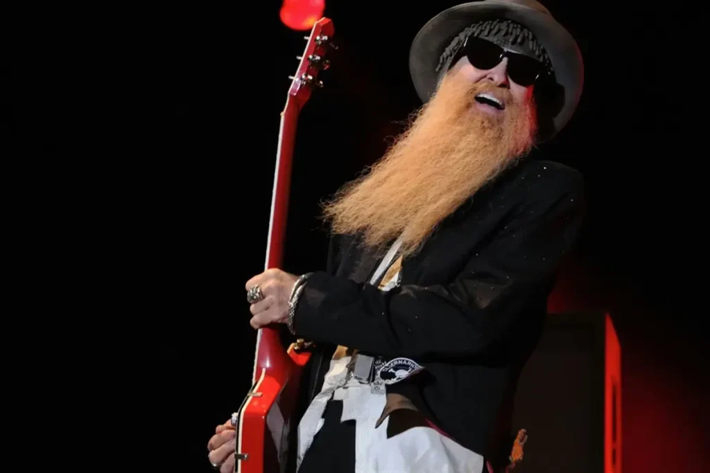 Billy Gibbons smiles mid-performance, gripping his guitar as his long beard and signature stage style stand out against a dark background.
