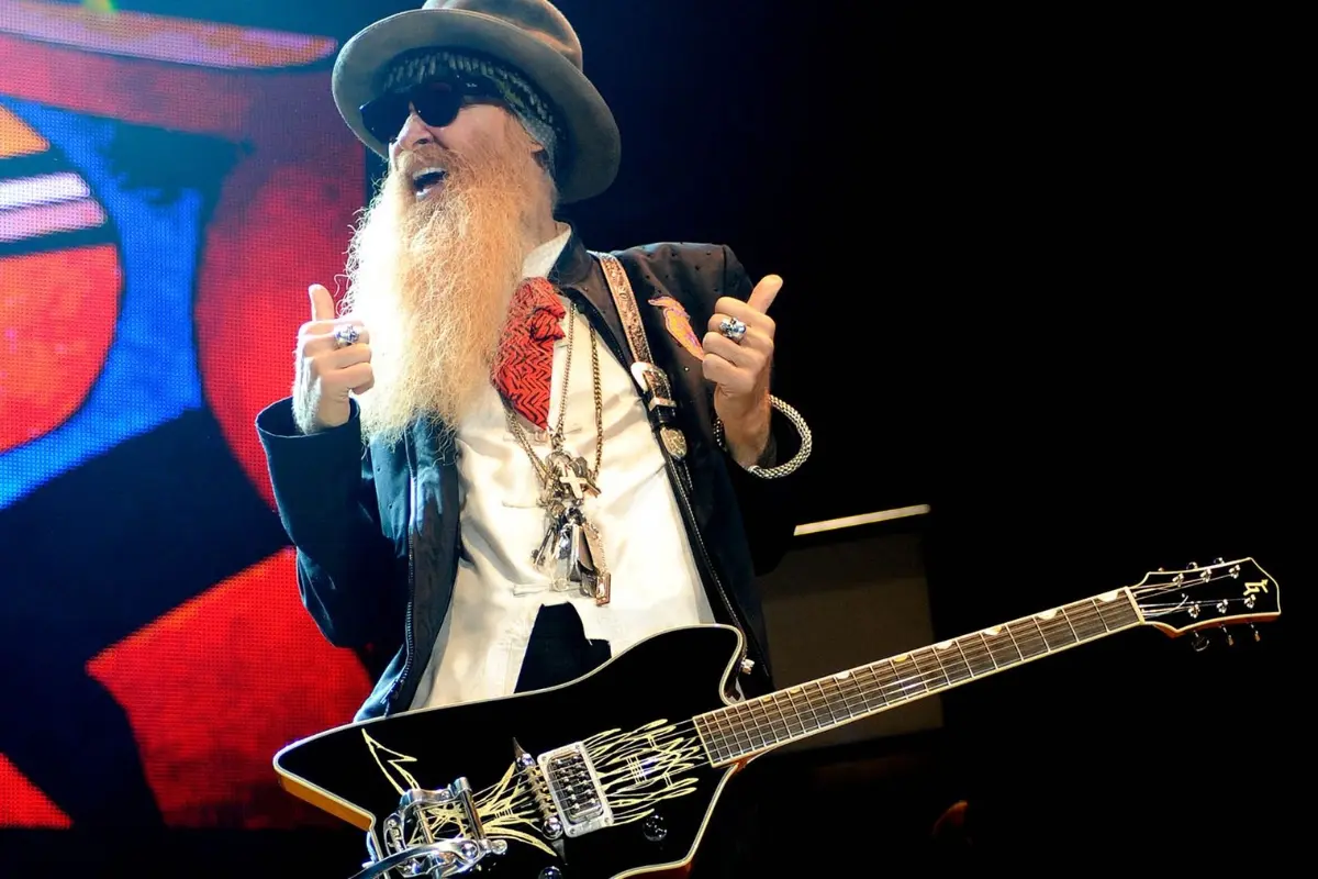 Billy Gibbons gestures to the crowd during a live show, electric guitar strapped across his chest with bold stage visuals behind him.