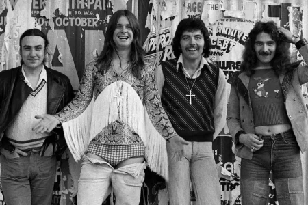 Black Sabbath band members posing casually in front of a wall covered with concert posters.