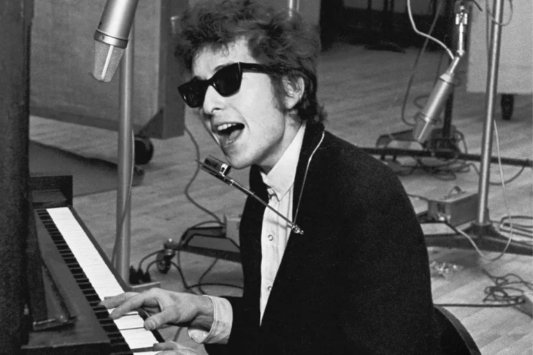 Bob Dylan wearing dark sunglasses seated at a piano.